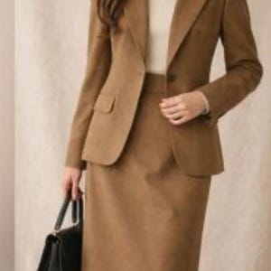 May include: A two-piece brown suit, consisting of a blazer and a knee-length skirt. The blazer has a notched lapel and two front pockets. A black handbag is held in the hand. The suit is paired with a white turtleneck. The outfit is suitable for business or formal occasions.