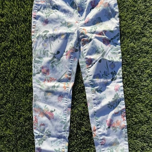 May include: Light-wash denim jeans with a colorful floral pattern. The jeans feature a high-waisted design and a classic five-pocket style. The floral print includes pink, purple, and green flowers and foliage.