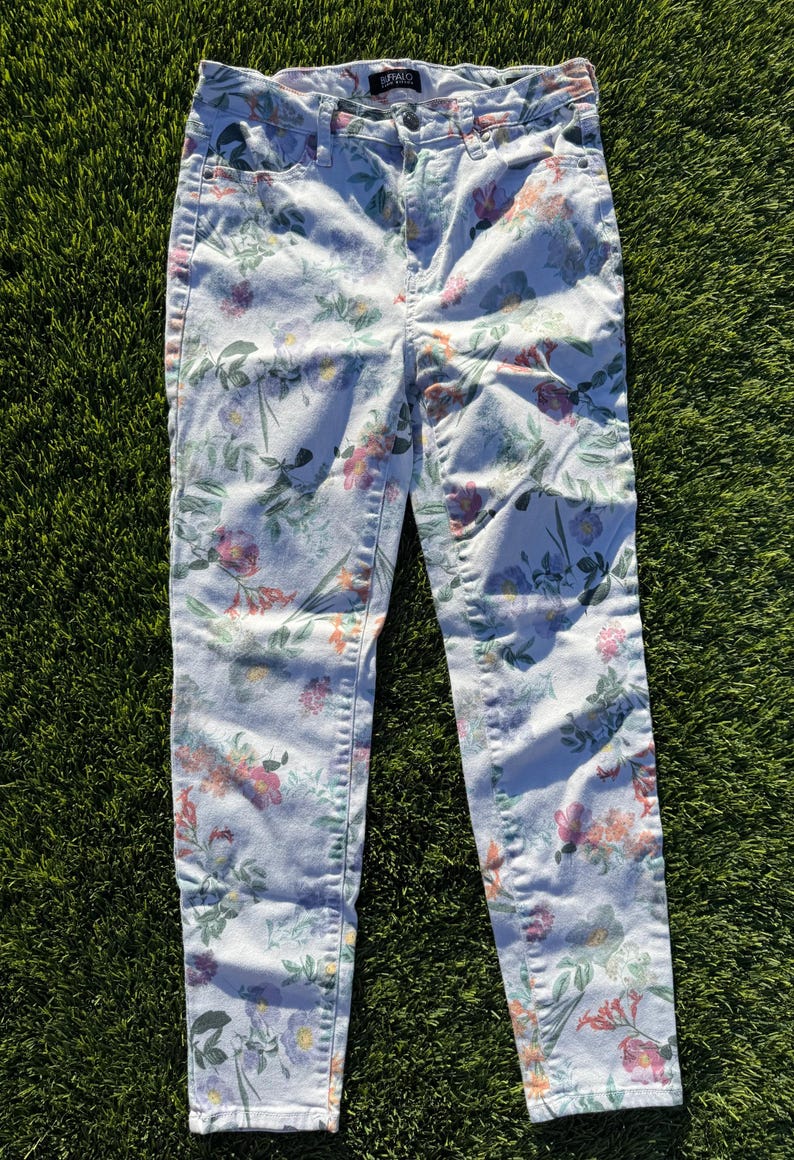 May include: Light-wash denim jeans with a colorful floral pattern. The jeans feature a high-waisted design and a classic five-pocket style. The floral print includes pink, purple, and green flowers and foliage.