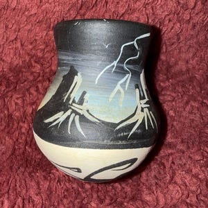 May include: A small, hand-painted ceramic vase with a black top and a cream-colored base. The vase features a landscape design with black mountains, white lightning bolts, and a blue and gray sky. The vase is approximately 4 inches tall.