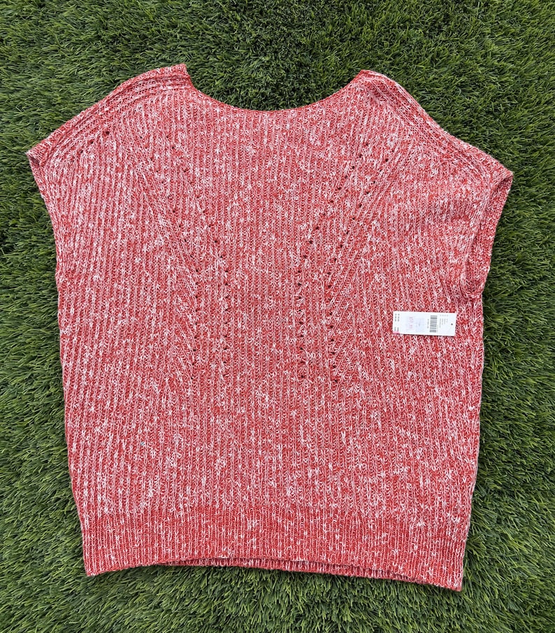 May include: Red and white knit sleeveless top. The top has a scoop neck and a textured knit pattern. The design includes a subtle V-shaped detail on the front. A tag is attached to the bottom right of the top.