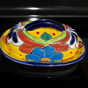 May include: A ceramic butter dish with a lid featuring a vibrant hand-painted floral pattern in yellow, blue, green, and red. The lid has a dark blue arched handle, and the base is rimmed with a matching dark blue border, creating a colorful design.