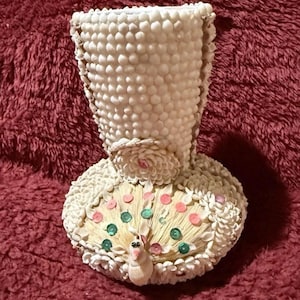 May include: A decorative vase made of white seashells. The vase has a flared top and a round base with a peacock design. The peacock's tail feathers are adorned with pink and green accents. The vase is approximately 7 inches tall.