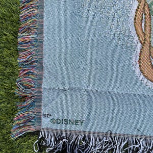 May include: Light blue woven throw blanket with a decorative design and fringed edges. The blanket has a textured appearance and features a multi-colored fringe on one side and a black and white fringe on the other. The word "&copy;DISNEY" is visible.