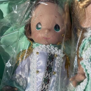 May include: Two dolls are visible, one in focus. The doll in focus has a smiling face with large, green eyes. It wears a white and light blue satin jacket with gold buttons and trim. The jacket has a decorative band with a geometric pattern. The doll is packaged in plastic.