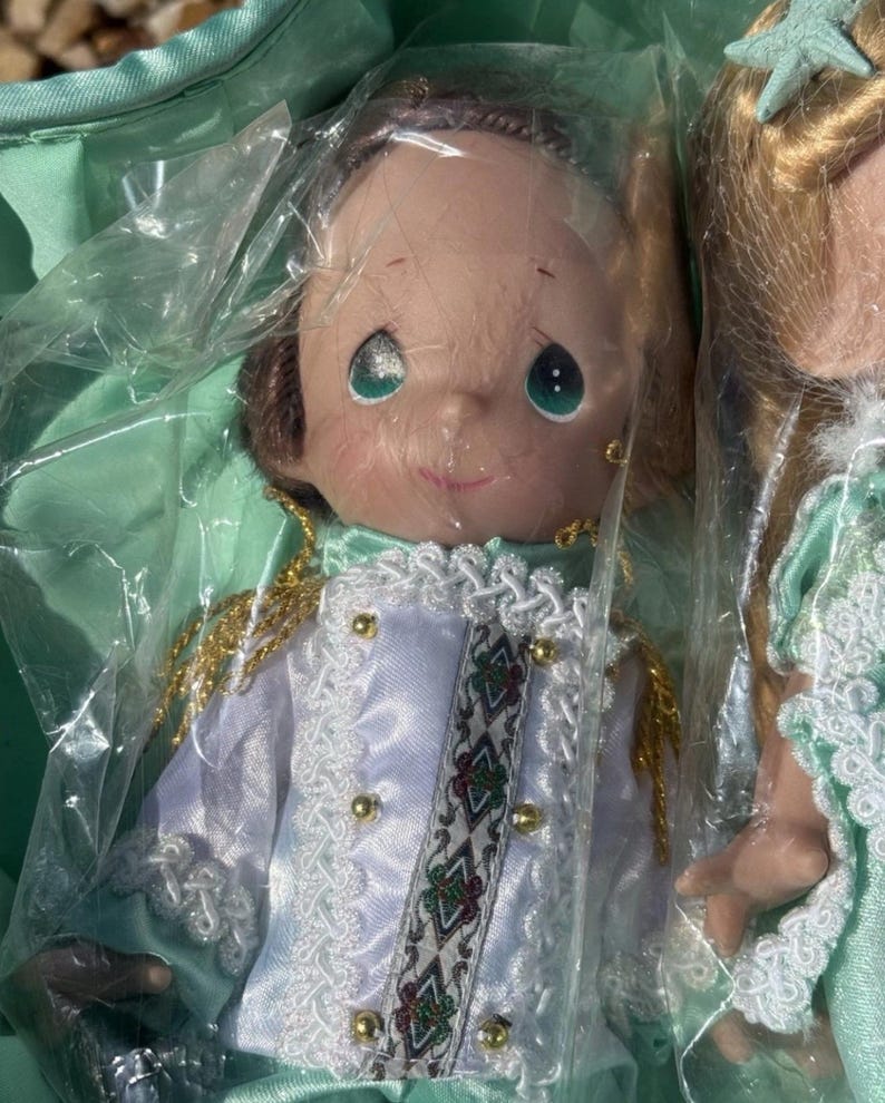 May include: Two dolls are visible, one in focus. The doll in focus has a smiling face with large, green eyes. It wears a white and light blue satin jacket with gold buttons and trim. The jacket has a decorative band with a geometric pattern. The doll is packaged in plastic.