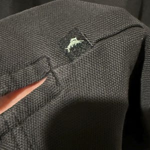 May include: Close-up of a dark gray polo shirt. The fabric has a textured weave. A small black tag with a white logo of a jumping fish is sewn onto the shirt.