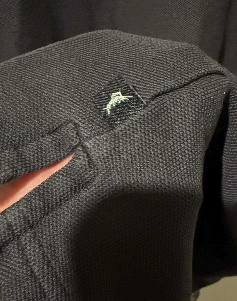 May include: Close-up of a dark gray polo shirt. The fabric has a textured weave. A small black tag with a white logo of a jumping fish is sewn onto the shirt.