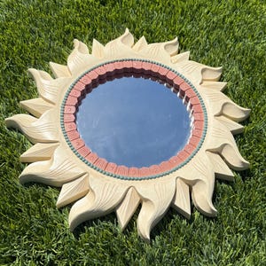 May include: A decorative sunburst mirror with a round reflective surface. The mirror is surrounded by a cream-colored frame with carved sun ray details. The inner ring features a coral-colored band with teal accents.