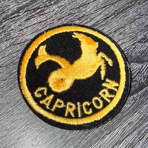 May include: A circular embroidered patch featuring a black background with a golden yellow border. The design depicts a stylized Capricorn sea-goat silhouette in golden yellow thread, with the word CAPRICORN stitched in yellow.