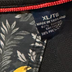 May include: Close-up of a navy blue garment tag with the text "XL/TG MADE IN THAILAND 62% COTTON 38% POLY". The tag is attached to a dark fabric with a floral pattern and red stitching.