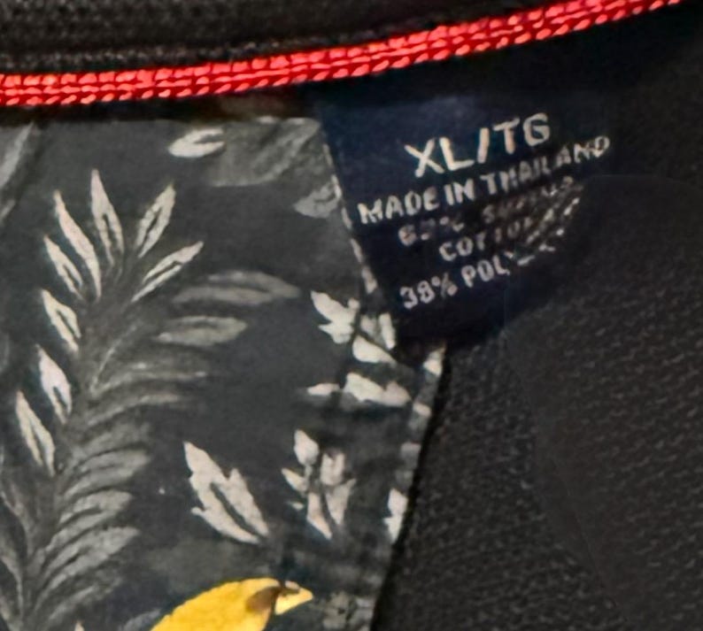 May include: Close-up of a navy blue garment tag with the text "XL/TG MADE IN THAILAND 62% COTTON 38% POLY". The tag is attached to a dark fabric with a floral pattern and red stitching.