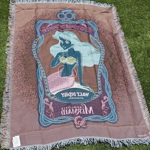 May include: A woven throw blanket featuring an image of Ariel from Disney's The Little Mermaid. The blanket has a brown background with a framed image of Ariel in blue, green, and pink. The blanket has a fringed edge.