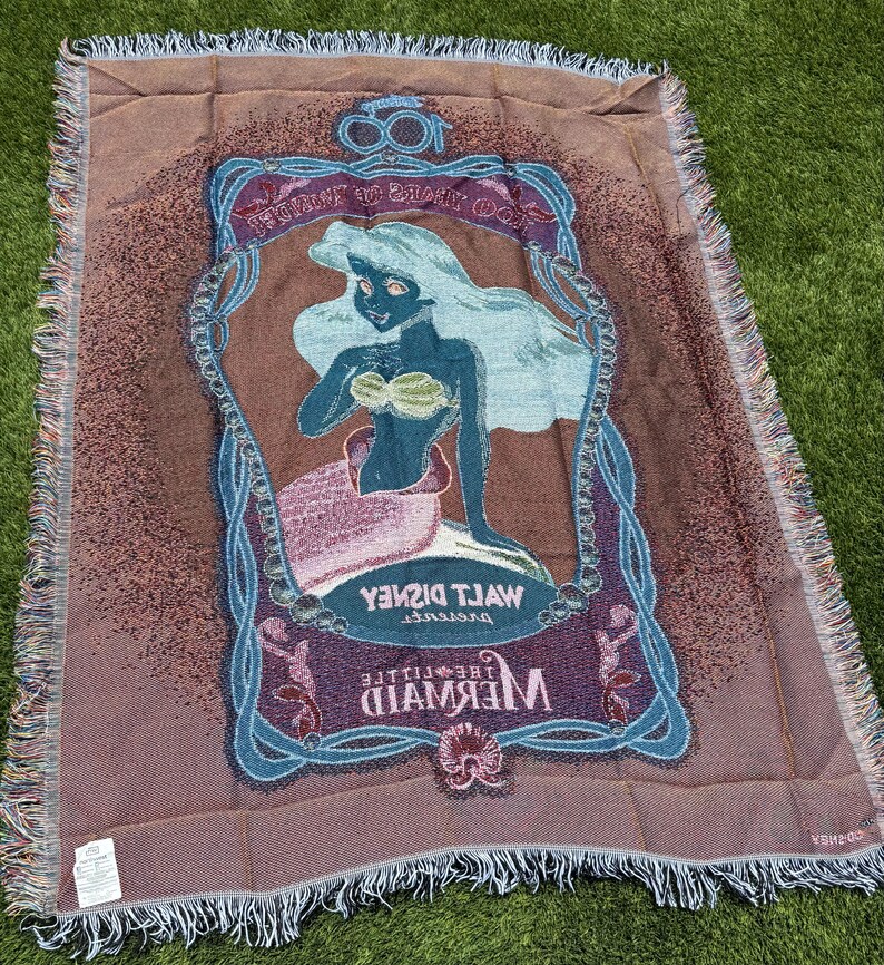 May include: A woven throw blanket featuring an image of Ariel from Disney's The Little Mermaid. The blanket has a brown background with a framed image of Ariel in blue, green, and pink. The blanket has a fringed edge.