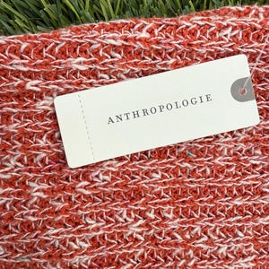 May include: Close-up of a knitted textile in shades of red and white. The fabric has a textured appearance. A white tag with the word "ANTHROPOLOGIE" is attached to the textile.
