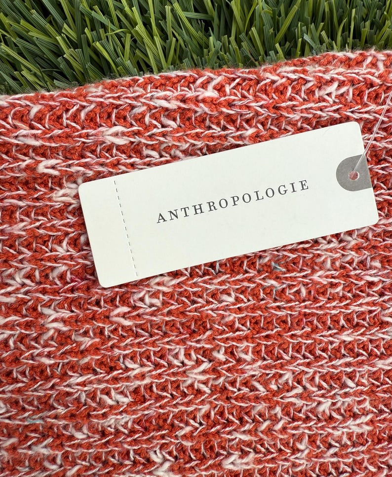 May include: Close-up of a knitted textile in shades of red and white. The fabric has a textured appearance. A white tag with the word "ANTHROPOLOGIE" is attached to the textile.