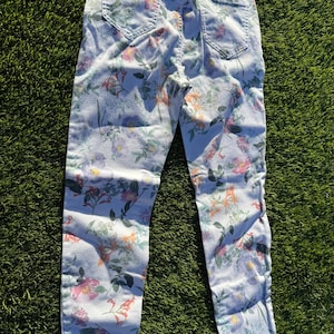 May include: Light blue denim jeans with a vibrant floral pattern. The jeans feature a mix of pink, orange, and green flowers and foliage. The jeans are laid flat on a green surface.