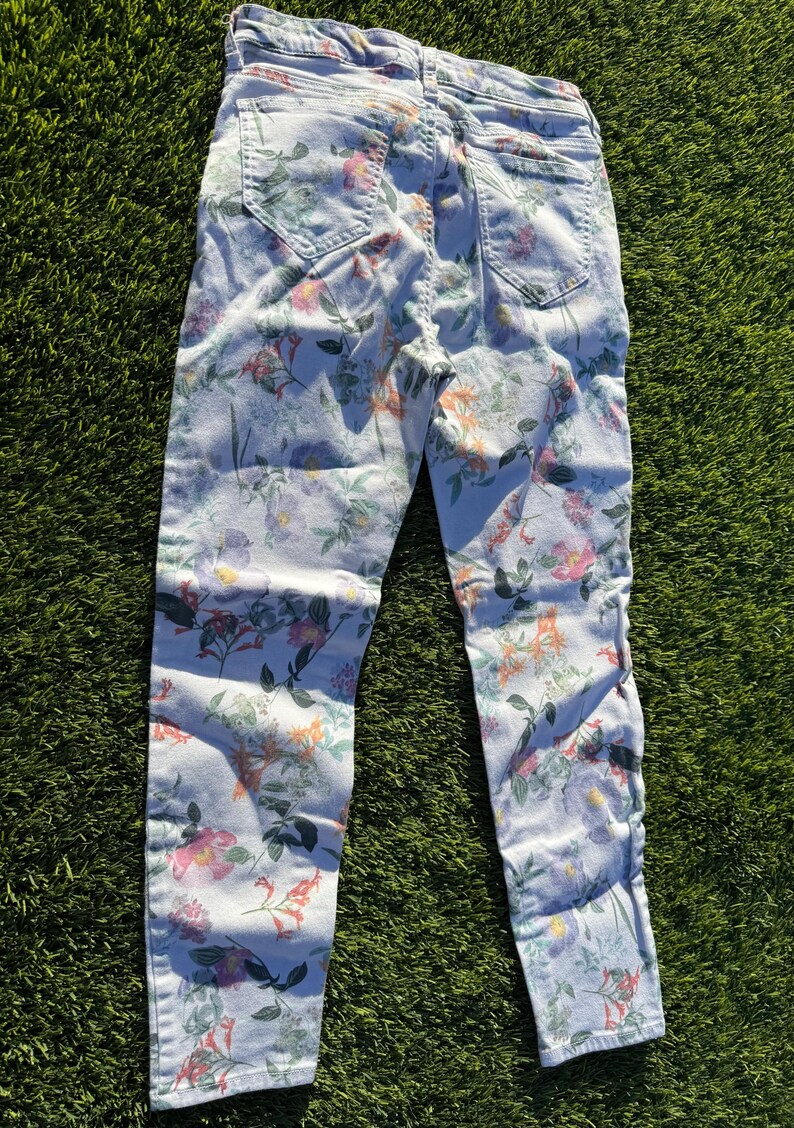 May include: Light blue denim jeans with a vibrant floral pattern. The jeans feature a mix of pink, orange, and green flowers and foliage. The jeans are laid flat on a green surface.
