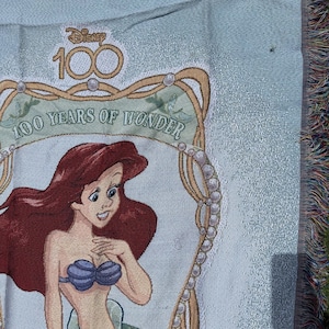 May include: Light blue woven throw blanket featuring Ariel from The Little Mermaid. The blanket has a decorative border with the text "100 Years of Wonder" and the Disney logo. The blanket has a fringed edge.