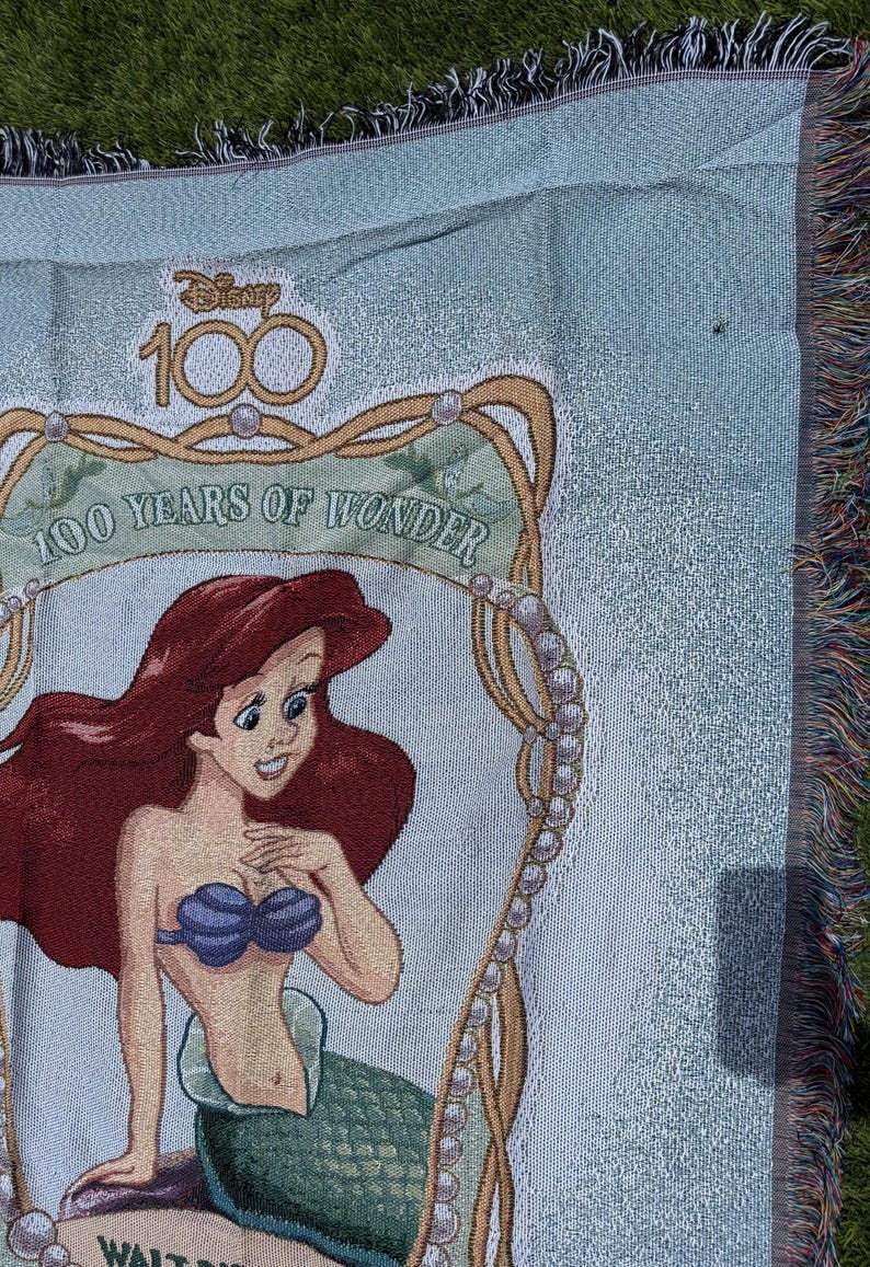 May include: Light blue woven throw blanket featuring Ariel from The Little Mermaid. The blanket has a decorative border with the text "100 Years of Wonder" and the Disney logo. The blanket has a fringed edge.