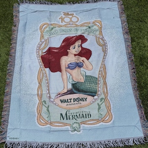 May include: Light blue throw blanket featuring Ariel from Disney's The Little Mermaid. The blanket has a fringed edge and includes the text "100 Years of Wonder" and "Walt Disney Presents The Little Mermaid".