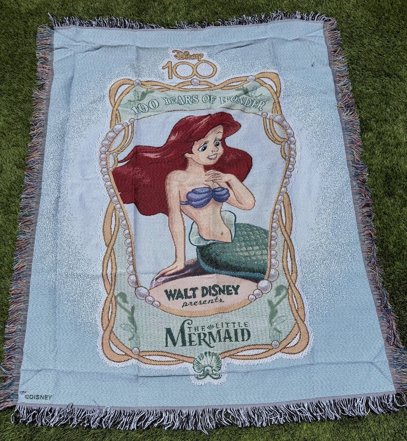 May include: Light blue throw blanket featuring Ariel from Disney's The Little Mermaid. The blanket has a fringed edge and includes the text "100 Years of Wonder" and "Walt Disney Presents The Little Mermaid".