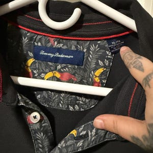 May include: Black polo shirt with a patterned collar featuring toucans and foliage. The shirt has a button-down collar and the Tommy Bahama logo. The size tag reads XL/TG.