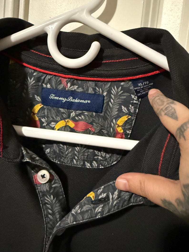 May include: Black polo shirt with a patterned collar featuring toucans and foliage. The shirt has a button-down collar and the Tommy Bahama logo. The size tag reads XL/TG.