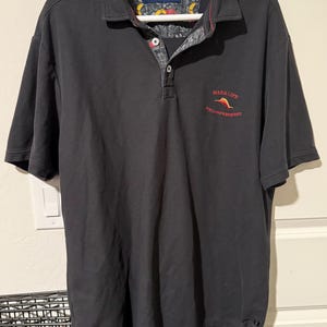May include: Black polo shirt with a collared neckline and short sleeves. The shirt features a floral pattern on the inner collar and the text "MAKE LIFE A WEEKEND" in red. The shirt is hanging on a white hanger.