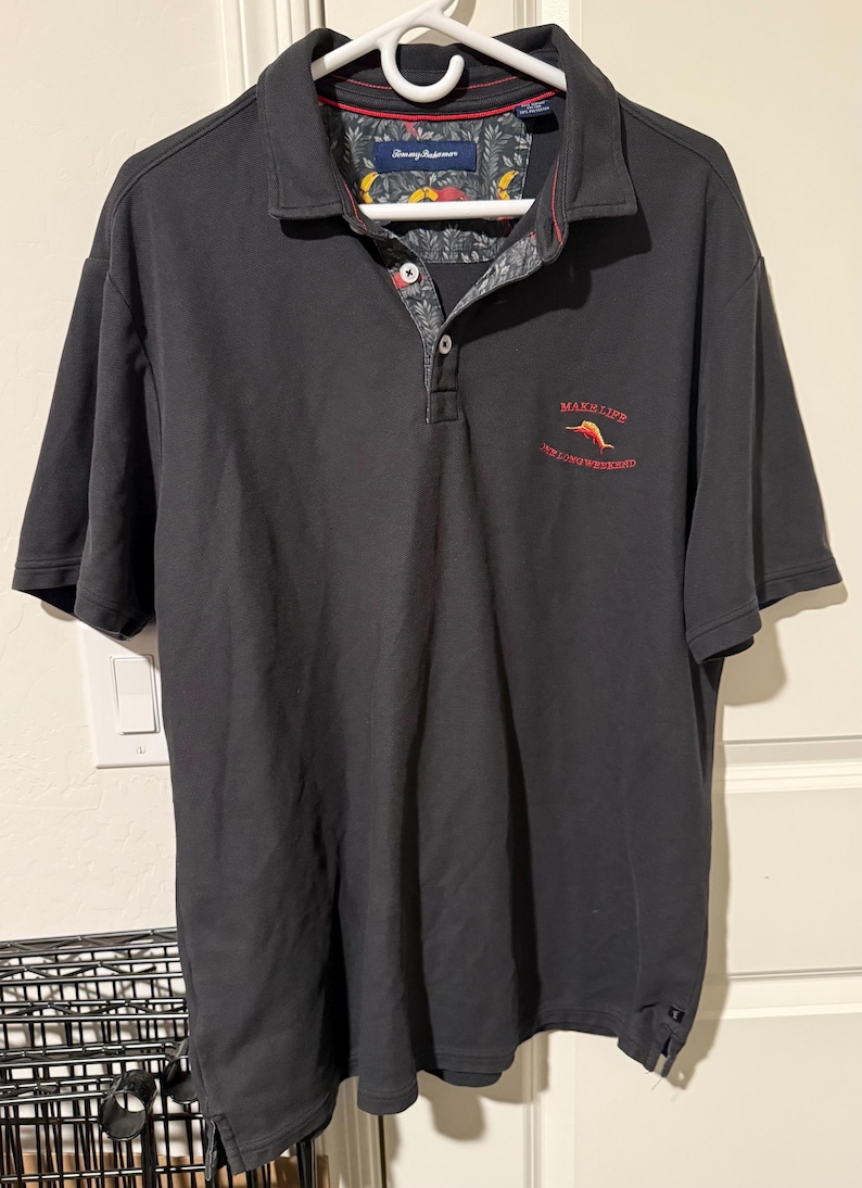 May include: Black polo shirt with a collared neckline and short sleeves. The shirt features a floral pattern on the inner collar and the text "MAKE LIFE A WEEKEND" in red. The shirt is hanging on a white hanger.