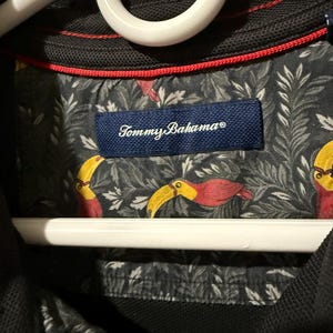 May include: Black polo shirt with a red trim and a patterned interior featuring toucans and foliage. The shirt has a navy blue label with the brand name "Tommy Bahama". The shirt is hanging on a white hanger.