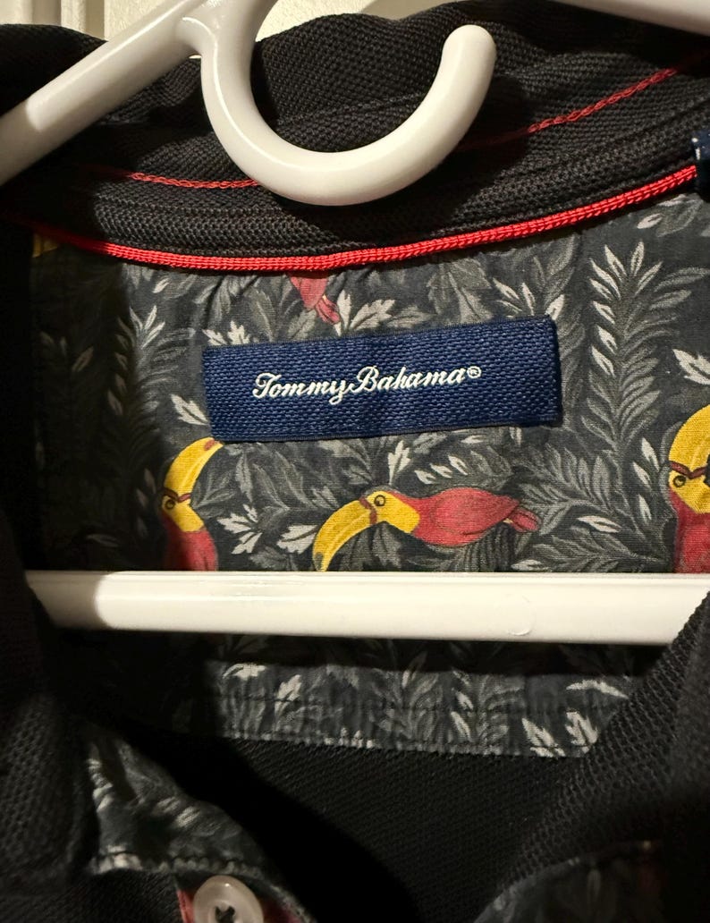 May include: Black polo shirt with a red trim and a patterned interior featuring toucans and foliage. The shirt has a navy blue label with the brand name "Tommy Bahama". The shirt is hanging on a white hanger.