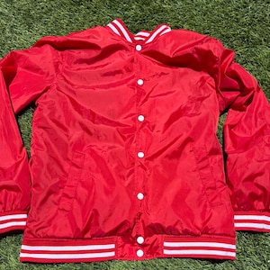 May include: A red satin jacket with white and red striped trim on the collar, cuffs, and waist. The jacket has white buttons down the front and two front pockets. The jacket is laying on a green surface.