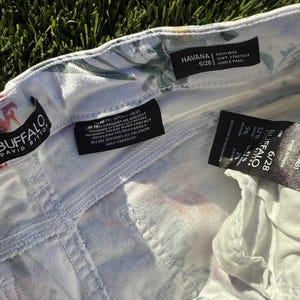 May include: Close-up of white denim ankle pants with a floral pattern. The pants have a high-rise fit and soft stretch. The brand label reads "BUFFALO DAVID BITTON". The size is 6/28.