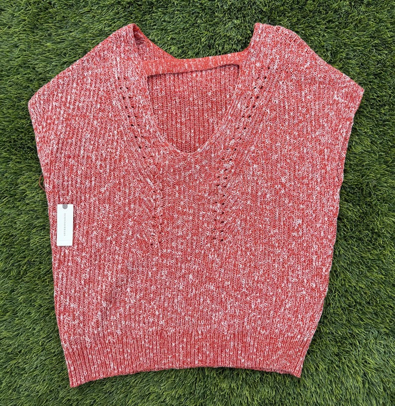 May include: A sleeveless red and white speckled knit sweater with a V-neck and a small cutout at the back of the neck. The sweater has a tag attached to the side. The sweater is made of a soft, comfortable material.