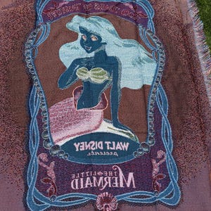 May include: A woven throw blanket featuring an image of Ariel from Disney's "The Little Mermaid." The blanket has a brown background with a blue and purple border. The image of Ariel is in the center, with the words "Walt Disney" and "The Little Mermaid" displayed.