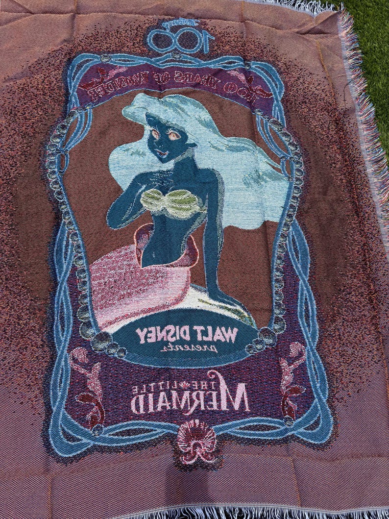 May include: A woven throw blanket featuring an image of Ariel from Disney's "The Little Mermaid." The blanket has a brown background with a blue and purple border. The image of Ariel is in the center, with the words "Walt Disney" and "The Little Mermaid" displayed.