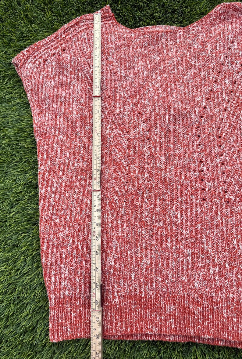 May include: Red and white knit sweater with a V-neck design. The sweater is approximately 27 inches long, measured with a wooden ruler. The fabric has a textured appearance with small holes throughout the design. The sweater is sleeveless.