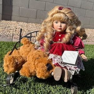May include: A porcelain doll with blonde curly hair and a red velvet dress, seated on a black metal carriage with a brown plush dog. The doll has a Certificate of Authenticity tag.