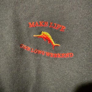 May include: Close-up of a dark gray polo shirt with red embroidered text that reads "MAKE LIFE" above "ONE LONG WEEKEND". A yellow and red dolphin is embroidered below "MAKE LIFE".