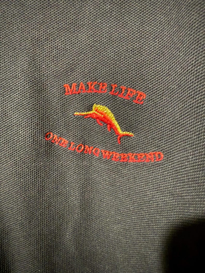 May include: Close-up of a dark gray polo shirt with red embroidered text that reads "MAKE LIFE" above "ONE LONG WEEKEND". A yellow and red dolphin is embroidered below "MAKE LIFE".