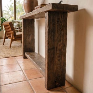 May include: A tall, narrow console table constructed from dark, weathered reclaimed wood with a visible grain pattern. The piece features a thick rectangular top, two sturdy vertical side supports, and a lower shelf connecting the legs.