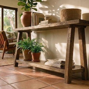 May include: A rectangular wooden console table with a lower shelf, featuring a weathered brown finish and four angled legs. The top surface holds a terracotta pot, books, and a woven basket, while the lower shelf holds two potted plants.