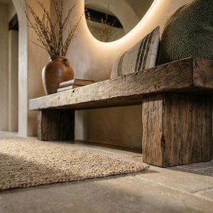 May include: A low, rectangular bench crafted from thick, weathered dark brown wood with a prominent grain. The bench features a sturdy, block-style base and is topped with a striped cushion and a textured green pillow.
