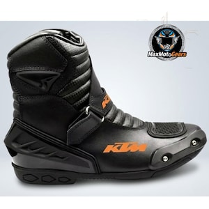 Könnte beinhalten: Schwarzer Motorradstiefel mit orangefarbenem KTM-Logo. Der Stiefel hat ein hohes Design mit verstärkten Einsätzen, einem Schnallenverschluss und einer strukturierten Sohle. Für Schutz und Leistung konzipiert.