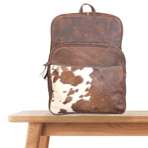 Puede incluir: Una mochila de cuero marrón con un bolsillo frontal de piel de vaca. La mochila tiene un asa superior y un bolsillo frontal con cremallera y una borla. El estampado de piel de vaca es marrón y blanco.