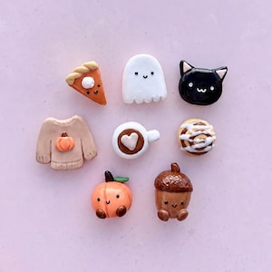 Puede incluir: Una colección de ocho pequeños dijes de arcilla de colores que representan una rebanada de pastel de calabaza, un fantasma blanco, una cara de gato negro, un suéter beige con una calabaza, una taza de café con un corazón, un rollo de canela, un melocotón y una bellota, todos con caras.