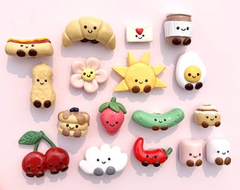 Può includere: Una collezione di piccoli ciondoli in argilla colorata con varie forme di cibo e natura, tra cui un croissant, una fragola, una ciliegia, un sole, un fiore, un'arachide, un hot dog, un cetriolino, una tazza di caffè, una busta, una nuvola e marshmallow.