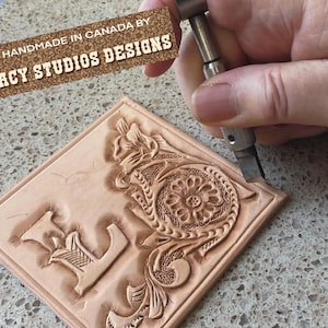 May include: A square piece of light brown leather with an embossed floral design and the letter "E". The text "HANDMADE IN CANADA BY STACY STUDIOS DESIGNS" is visible. A hand is using a tool to carve the leather.