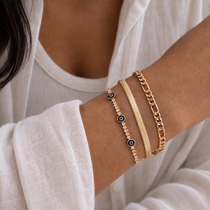 May include: A set of three gold-toned bracelets worn together. One bracelet features a delicate chain with small, round, blue and white evil eye charms. The second is a flat, flexible herringbone chain. The third is a classic gold link chain.